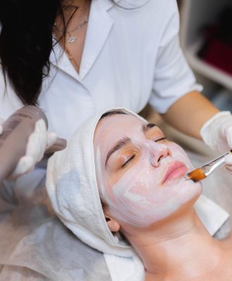 Beautician with a brush applies a white moisturizing mask to the face of a young girl client in a spa beauty salon